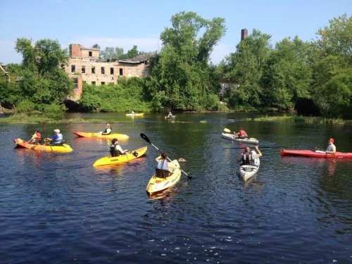 A group of kayakers paddles on a calm river, surrounded by lush greenery and a partially ruined building in the background.