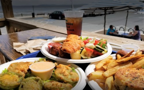 A table with two plates of fried food, a salad, and a drink, with a scenic view of the water in the background.