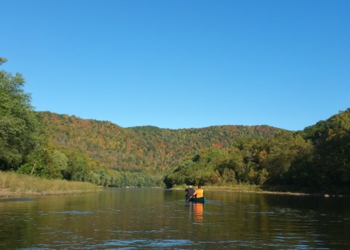 A serene river scene with two people kayaking, surrounded by colorful autumn foliage and clear blue skies.