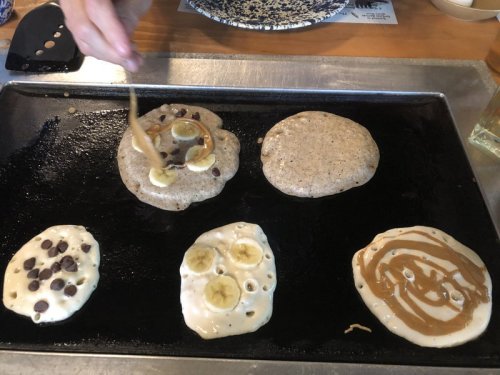 A hand drizzles batter onto pancakes with chocolate chips and banana slices on a griddle.
