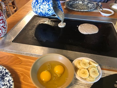 A griddle with a pancake cooking, alongside a bowl of eggs and sliced bananas on a wooden table.