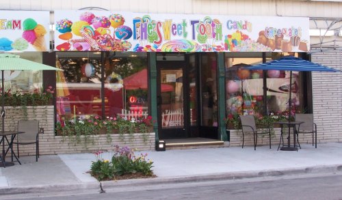 Colorful storefront of a candy shop with a sign reading "Fly's Sweet Tooth," featuring outdoor seating and decorations.
