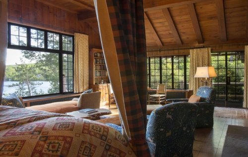 Cozy wooden living room with large windows overlooking a lake, featuring patterned furniture and warm lighting.