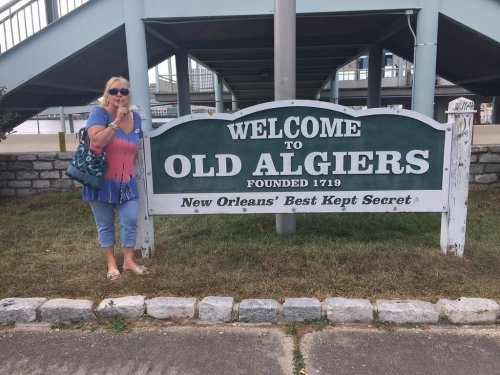 A woman stands beside a sign that reads "Welcome to Old Algiers, Founded 1719, New Orleans' Best Kept Secret."