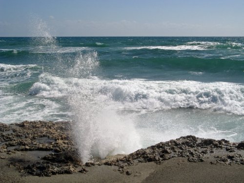 Waves crashing against rocky shore, creating a spray of water under a clear blue sky.