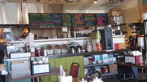 A cozy café interior with a chalkboard menu, coffee machines, and tables set for dining. Staff are preparing food.
