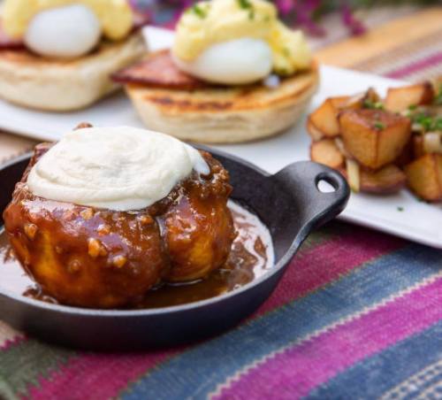 A skillet with a dessert featuring dough balls in sauce, topped with cream, alongside a plate of eggs and potatoes.