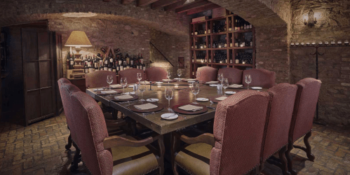A cozy, dimly lit dining room with a large wooden table surrounded by upholstered chairs and wine bottles on shelves.