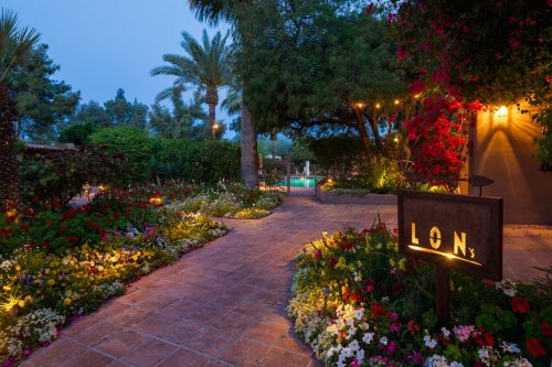 A beautifully lit garden path with vibrant flowers, leading to a pool area, surrounded by palm trees and greenery.
