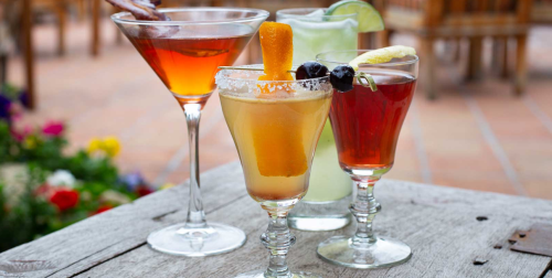 Three colorful cocktails on a wooden table, surrounded by flowers. A martini, a citrus drink, and a fruity beverage.
