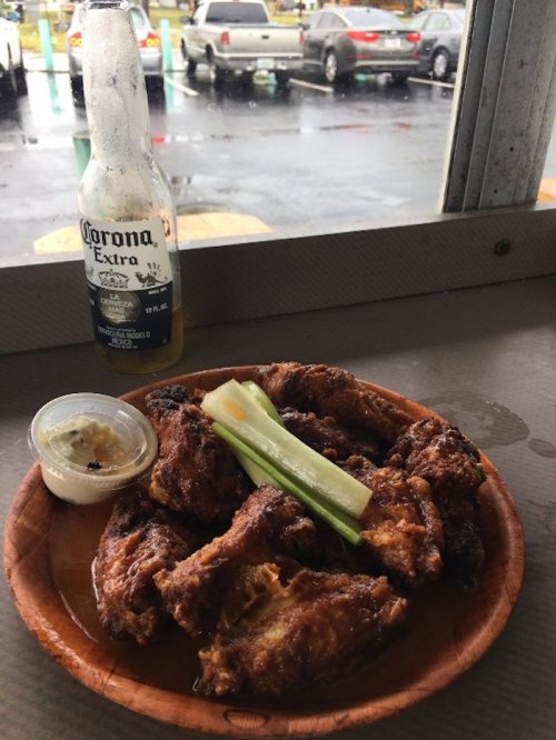 A wooden plate with crispy chicken wings, celery sticks, and a dipping sauce, alongside a bottle of Corona beer.
