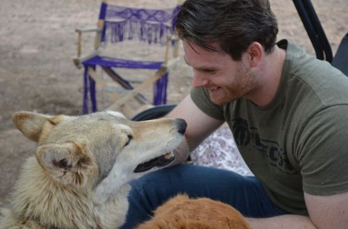 A man smiles while interacting with a friendly dog in a relaxed outdoor setting.