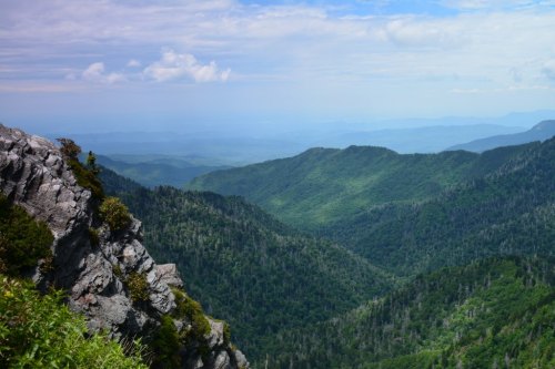 A panoramic view of lush green mountains under a partly cloudy sky, with rocky cliffs in the foreground.
