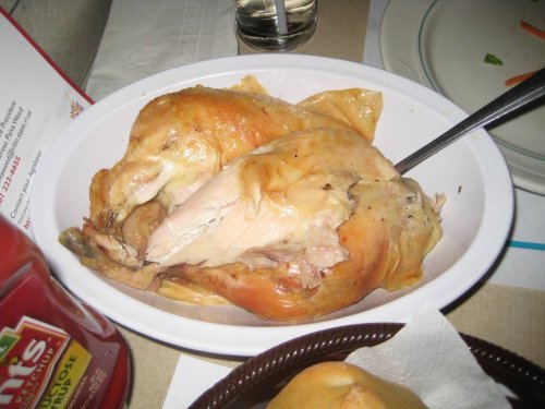A serving of roasted chicken on a white plate, with a spoon resting on top, accompanied by condiments in the background.