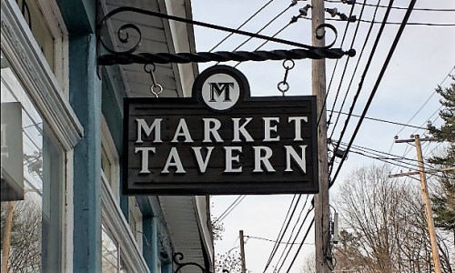 Sign for "Market Tavern" hanging from a building, with power lines and trees in the background.