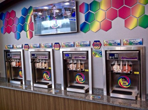 Four colorful ice cream machines with various flavors, displayed in a modern shop with a TV screen above.
