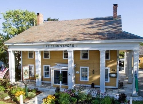 A historic yellow tavern with white columns, surrounded by greenery and flags, featuring a welcoming entrance.