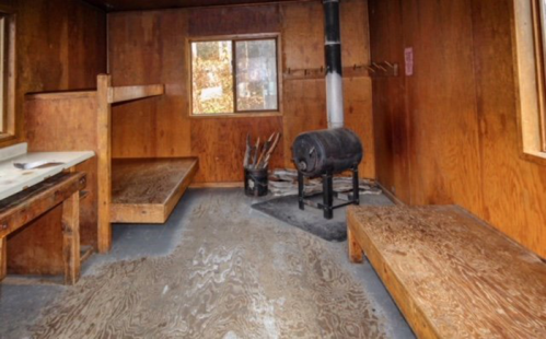 Interior of a rustic wooden cabin with two beds, a wood stove, and a window, featuring a simple, cozy design.