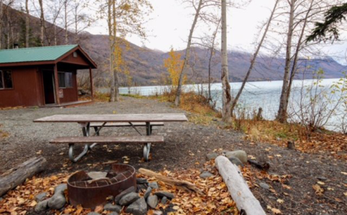 A picnic table near a fire pit by a lake, surrounded by autumn trees and mountains in the background.