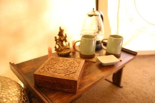 A wooden table with a carved box, two mugs, a small statue, and a kettle, set in a cozy, softly lit space.