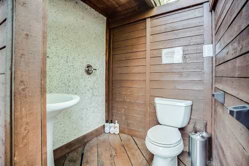 A rustic bathroom with wooden walls, a toilet, and a sink, featuring natural stone accents and toiletries on the floor.