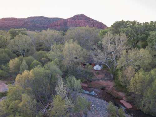 Aerial view of a tent nestled among lush trees by a stream, with red rock mountains in the background.