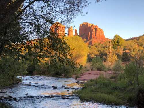 A serene river flows through a lush landscape, with red rock formations illuminated by the setting sun in the background.