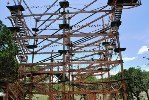 A large wooden climbing structure with ropes and platforms, surrounded by trees and blue sky.