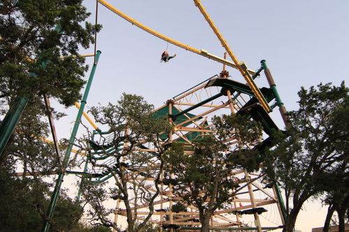 A person swings high above a colorful amusement park ride, surrounded by trees and a clear sky.