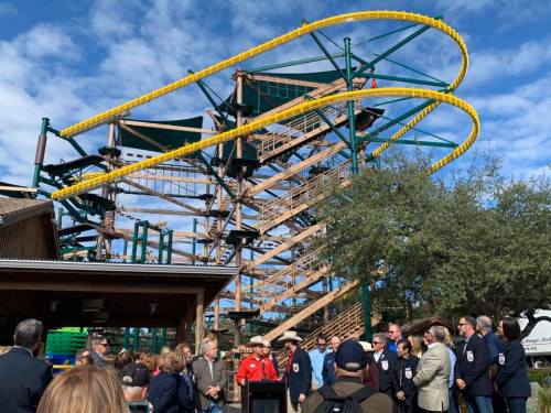 A large wooden roller coaster with yellow tracks towers over a crowd gathered for an event in front of it.