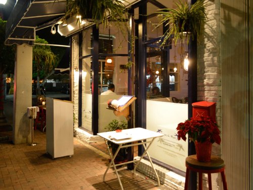 A cozy restaurant exterior at night, featuring plants, warm lighting, and a small table outside.