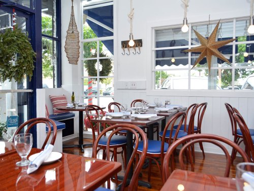 Bright restaurant interior with wooden tables, chairs, and nautical decor, featuring large windows and a cozy atmosphere.