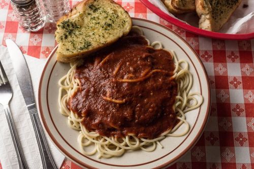 A plate of spaghetti topped with marinara sauce, accompanied by a slice of garlic bread on a checkered tablecloth.