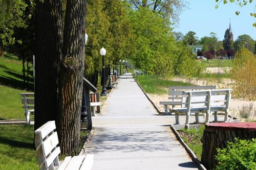 A peaceful park pathway lined with benches, trees, and greenery, leading towards a beach area in the distance.