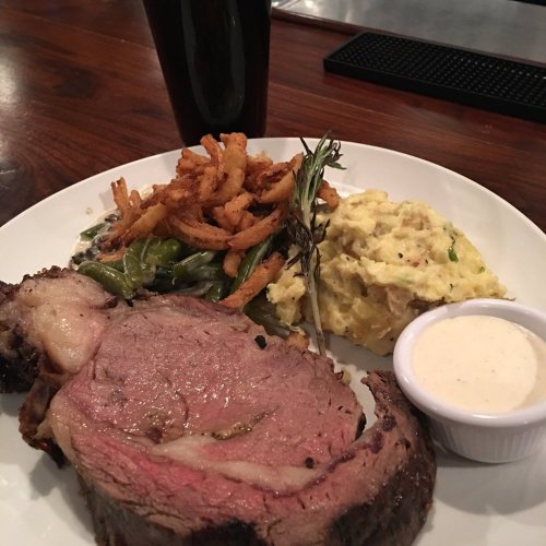 A plate featuring prime rib, mashed potatoes, green beans, crispy onions, and a dipping sauce, with a dark beverage.