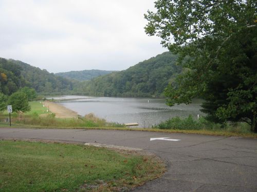 A serene lake surrounded by green hills under a cloudy sky, with a winding road leading to the water's edge.