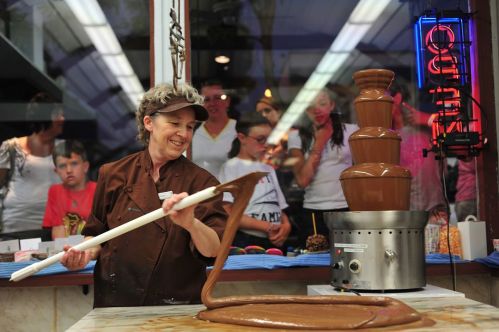 A chocolatier stirs melted chocolate near a fountain, with onlookers watching eagerly in the background.