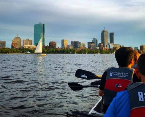 A group of people in a canoe on the Charles River, with a city skyline and sailboats in the background.
