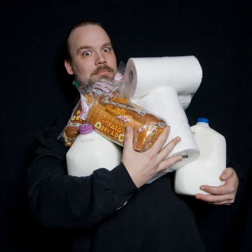 A person holds multiple items, including milk jugs, paper towels, and a loaf of bread, with a surprised expression.