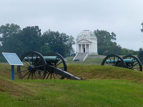 A historic site featuring cannons in the foreground and a monument on a hill in the background, under a cloudy sky.