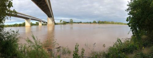 A panoramic view of a river under a bridge, with cloudy skies and lush greenery along the riverbank.