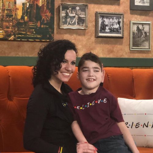 A woman and a boy sit together on a couch, smiling in a cozy, decorated room with framed photos on the walls.