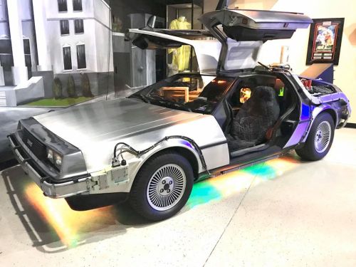A silver DeLorean car with gullwing doors, featuring colorful lights inside, displayed in a museum setting.