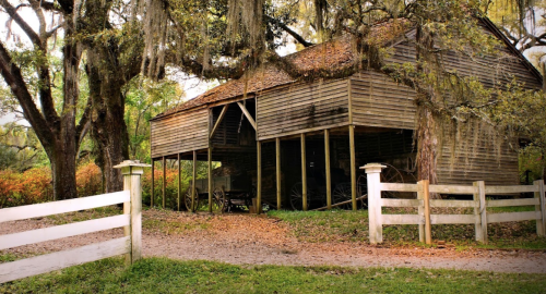A rustic wooden barn surrounded by trees, with a dirt path and white fences leading to it.