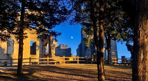 Ruins of a stone building framed by trees, with a clear blue sky and a visible moon in the background.