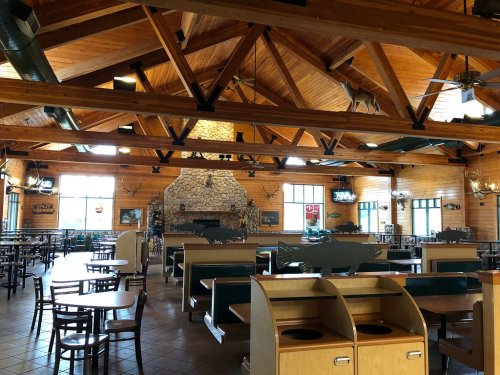 Interior of a rustic restaurant with wooden beams, stone fireplace, and empty tables. Large windows let in natural light.