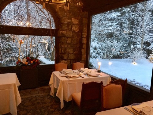 Cozy dining area with elegant tables, overlooking a snowy landscape through large windows. Warm ambiance with floral decor.
