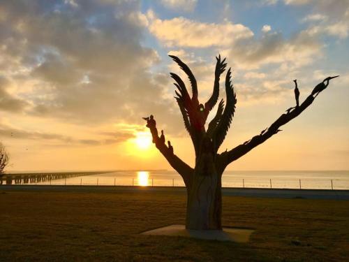 A wooden sculpture of a tree with bird figures, set against a sunset over the water and a pier in the background.