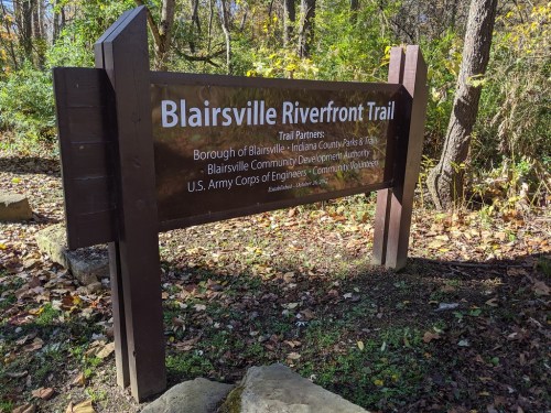 Sign for the Blairsville Riverfront Trail, listing partners and sponsors, surrounded by trees and autumn foliage.