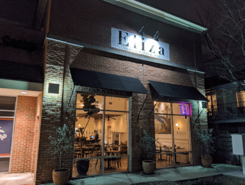 Exterior of a restaurant named "Eliza" at night, featuring large windows, outdoor seating, and illuminated signage.
